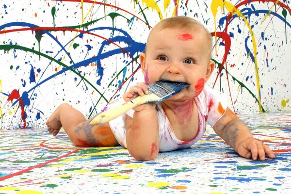 Un niño pequeño cubierto de pintura, lamiendo un pincel sonriente