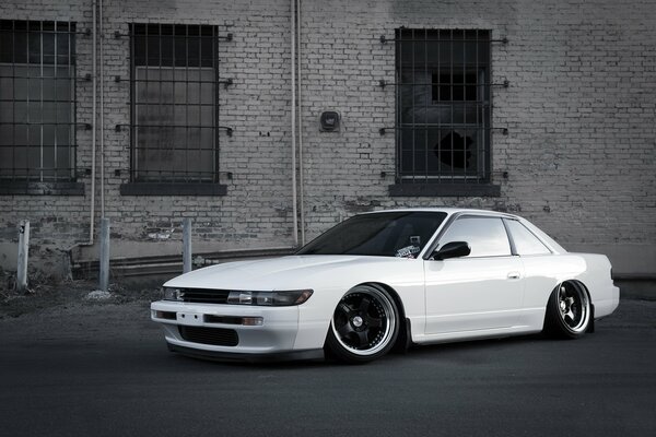 The car is a white nissan Sylvia s13 at a brick wall with windows in bars