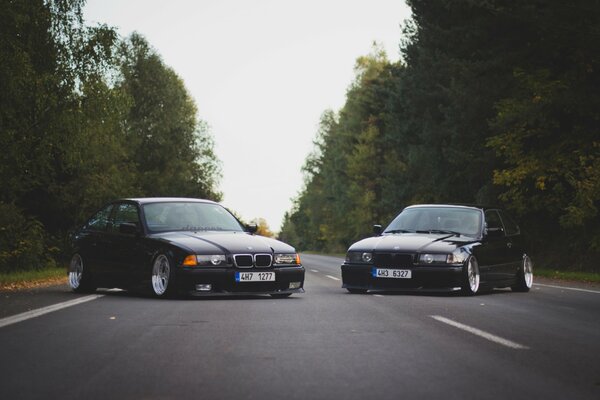 Автомобили BMW M3 e36 3 серии oldschool тюнингованные стоят поперек асфальтированной дороги на фоне леса вид спереди