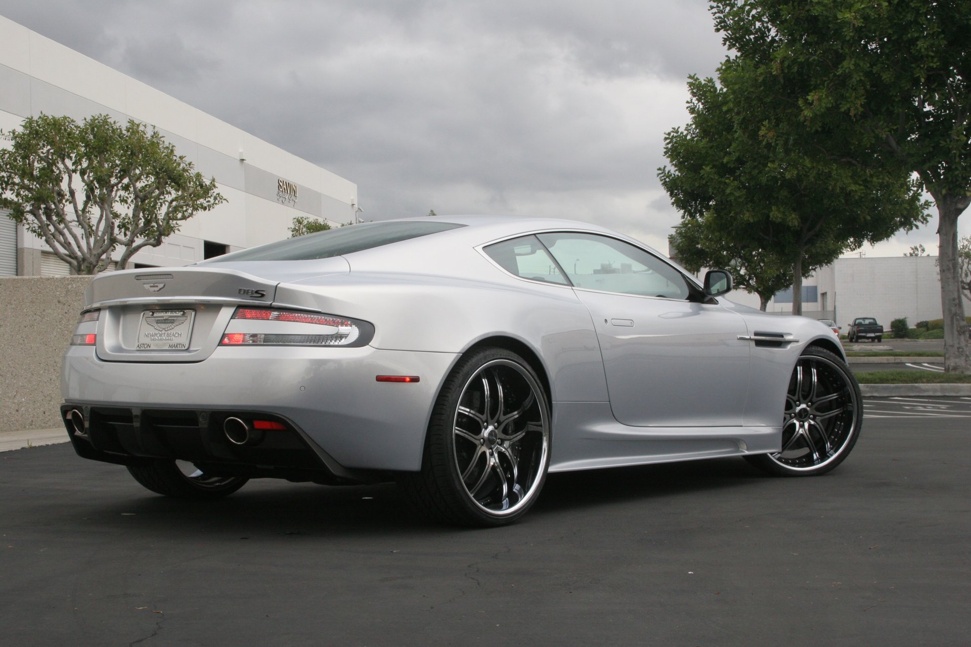 aston martin dbs silber räder aston martin dbs silber rückansicht scheinwerferlicht himmel wolken bäume gebäude