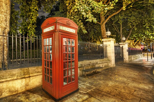 A red telephone booth at the fence of the park. Evening