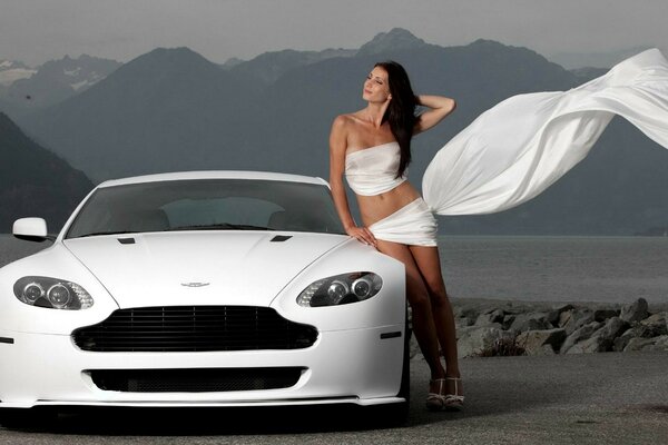 A white Aston Martin car. A girl in white near the car. A road on the background of mountains
