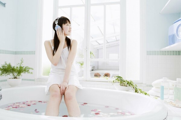 A young Asian woman with headphones is sitting in the bathtub