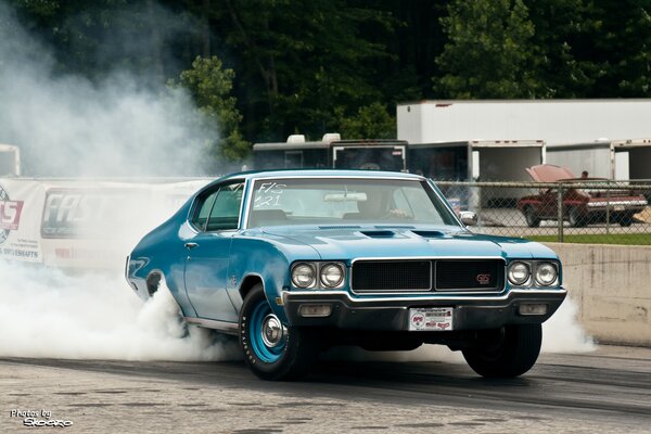 A blue car revving on the spot that has smoke coming out from under the wheels