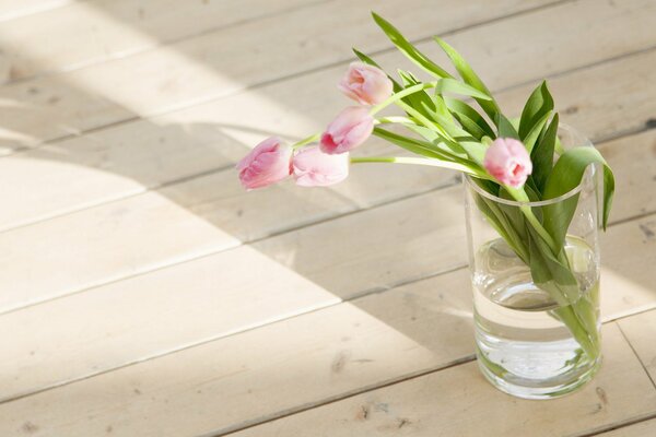 Tulips in a glass of water on a wooden floor in the rays of the spring morning sun