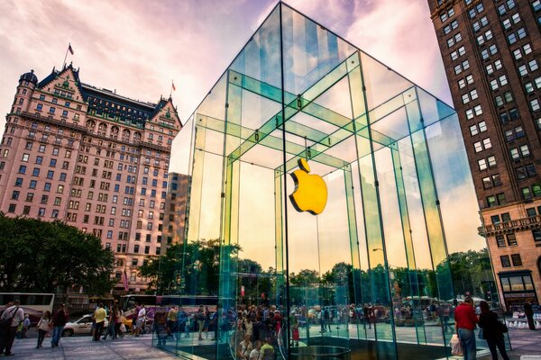 The street on which there are skyscrapers, people walk and in the center there is a glass structure with the apple logo