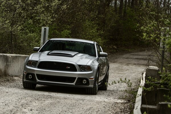 A silver Mustang car rides along the road outside the city