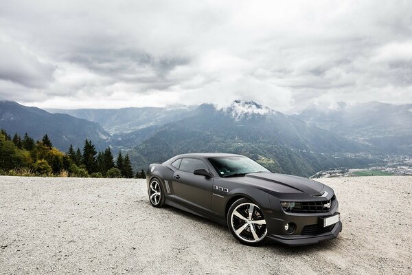 Coche negro Camaro Chevrolet en la carretera en medio de un bosque de montañas y cielo nublado