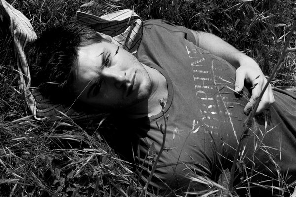 Un gars aux cheveux noirs pensif allongé sur l herbe. Beau mec aux cheveux noirs sur une photo en noir et blanc allongé dans l herbe