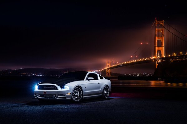 Im Vordergrund in der Nacht steht ein weißer Ford Mustang in der Nähe einer von Laternen beleuchteten Brücke, die den Blickwinkel von vorne angibt