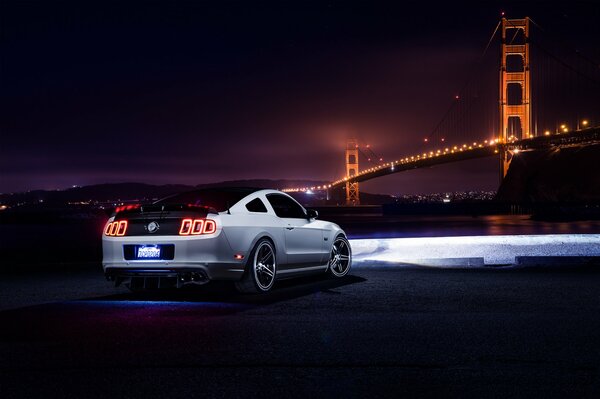 Ein weißer Ford Mustang in der Nacht, der den Weg zur Brücke mit Scheinwerfern beleuchtet, ein Blickwinkel von hinten