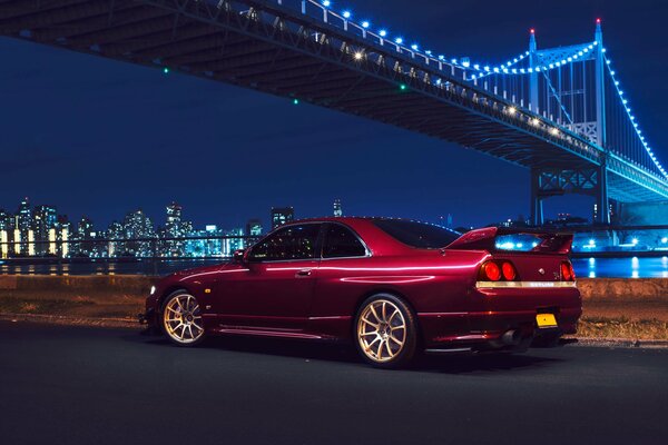 A red Nissan car stands under the New York Bridge at night