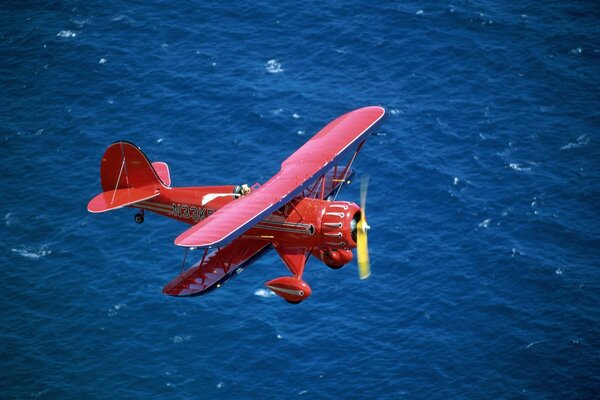 A red biplane flies over the sea