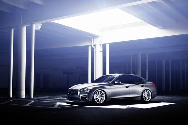 A metallic-colored Infiniti car under a bright window in the ceiling in an underground parking lot