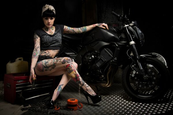 A girl with tattoos is sitting next to a black motorcycle