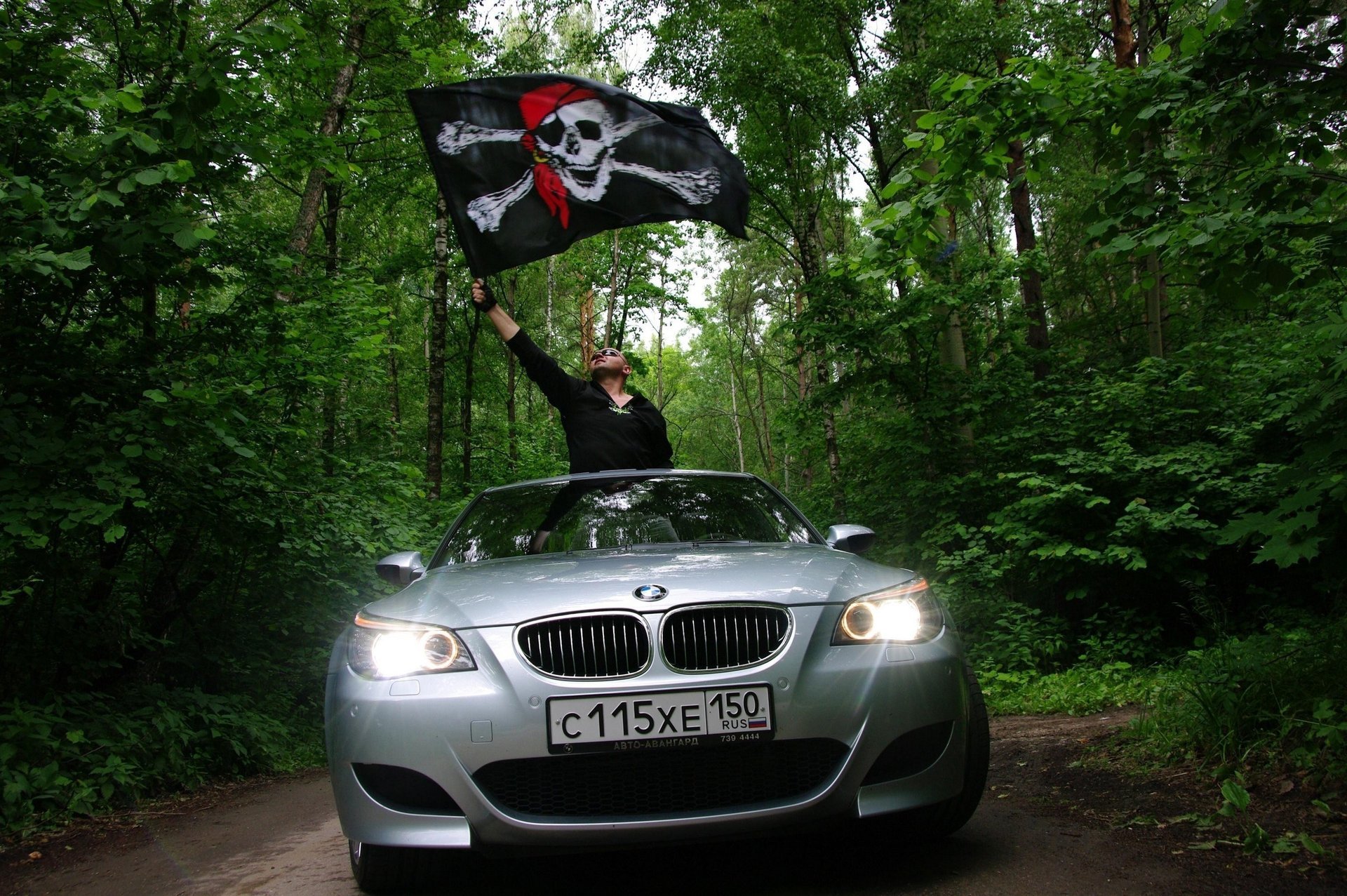 graue färbung mann mit flagge schwarze flagge schädel auto bmw grün wald natur lichter silbernes auto auto mann flagge pkw transport kraftfahrzeuge