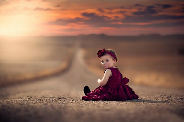 A little girl in a burgundy dress is sitting on the road