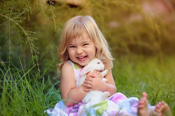 A joyful girl with a white bunny is sitting on the grass and laughing