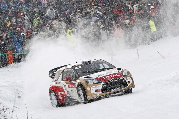 A car racing in snowy weather. The audience can be seen in the background