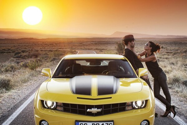 A couple on the road next to a Chevrolet on the background of sunset