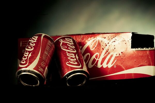 Due lattine di Coca Cola accanto alla scatola