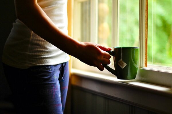 Eine grüne Tasse Tee in den Händen am Fenster