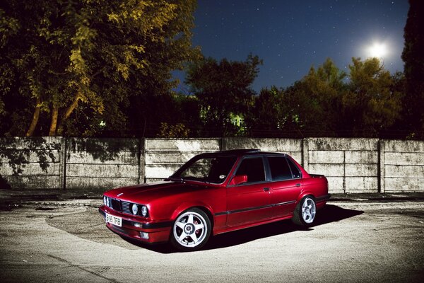 Czerwone BMW E30 na tle nocnego ogrodzenia przy księżycu
