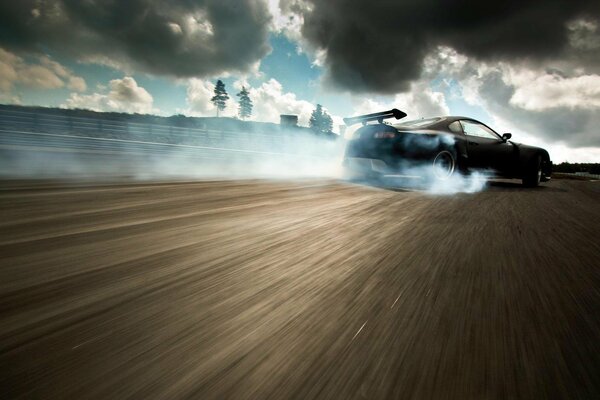 The drift of a black car against the background of a graphite highway and under a cloudy sky with black clouds of smoke