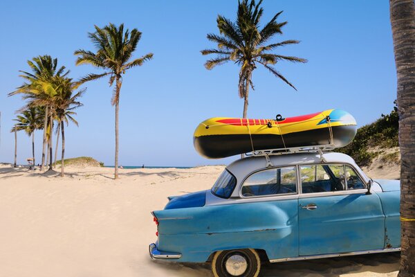 Soviet blue car with a boat on the roof on the sandy ocean shore under palm trees against the blue sky