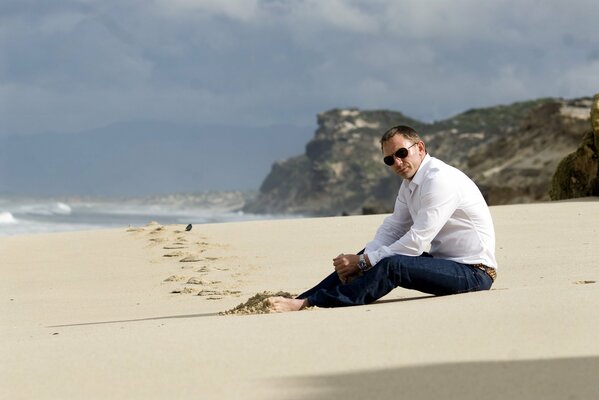 Daniel Craig in sunglasses sitting on the beach barefoot