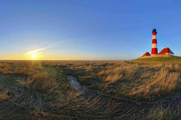 A panoramic morning opens up a view of a field of grass, a running stream and a lighthouse in the distance