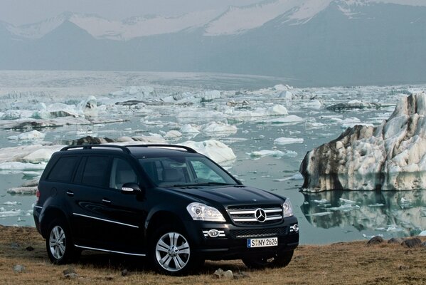 Mercedes SUV negro en la orilla de un cuerpo de agua con hielo derretido y montañas en la distancia