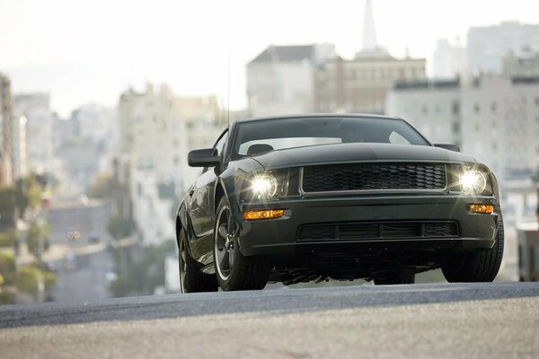 Ford mustang bullitt мчится по дороге на фоне городских многоэтажек