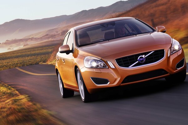 Coche Volvo naranja en la carretera en el campo en medio de las montañas y el cielo azul claro