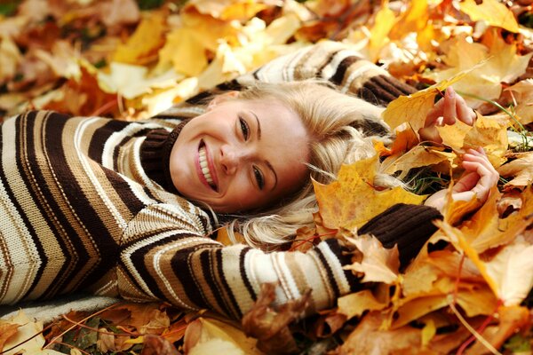 A blonde in a striped sweater lies on the fallen leaves of a maple tree