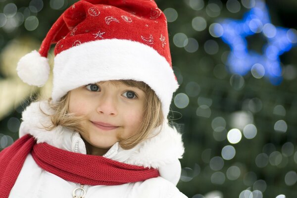 Chica con cara en un sombrero de año nuevo. Niña de ojos azules con sombrero de papá Noel. Foto de año nuevo con niños. Niña en el fondo del árbol de Navidad