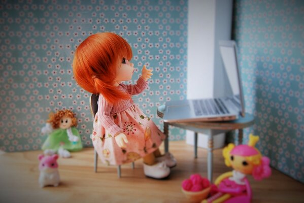 Muñeca con vestido rosa. Composición de muñecas. Muñeca detrás de la computadora portátil. La muñeca con el pelo de fuego y sus amigos