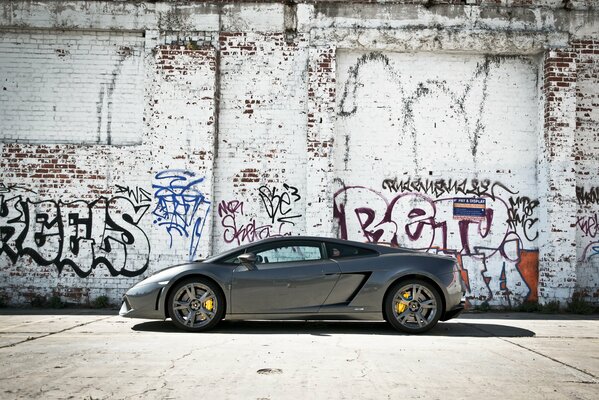 Серый автомобиль lamborghini gallardo на фоне стены с граффити