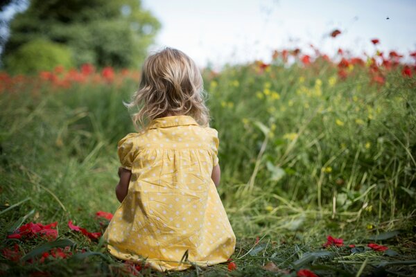 A little girl in a yellow summer sundress is sitting in a meadow