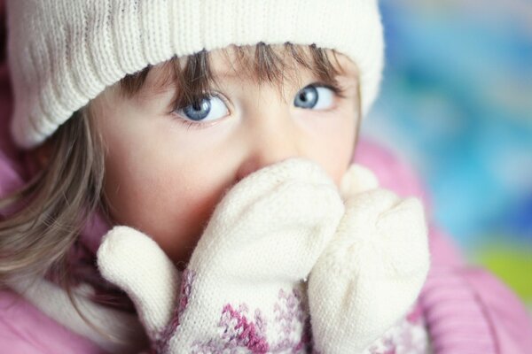 Ein blauäugiges Mädchen bedeckt ihr Gesicht mit ihren Händen in warmen weißen Handschuhen