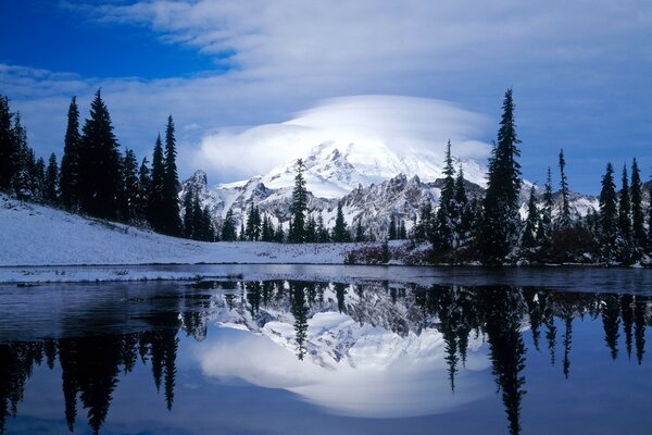 Volcán cubierto de nieve en las nubes y los árboles se reflejan en el lago