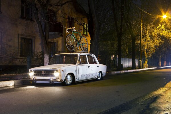 A white Lada with a bicycle on the roof is waiting for the owner at night