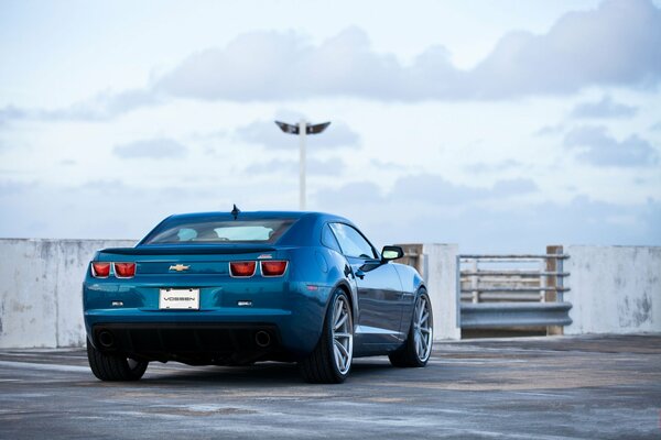 Une chevrolet Camaro SS bleue dans un parking au milieu d un ciel blanc sur lequel flottent des nuages blancs