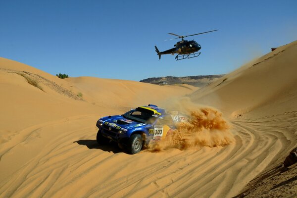 Niebieski SUV jedzie po piasku za nim leci helikopter