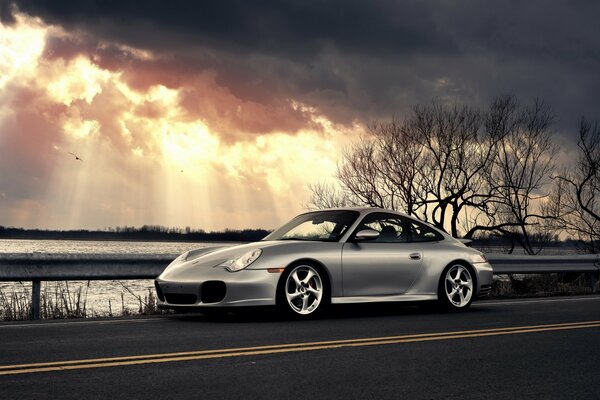 A silver Porsche stands next to the water under the clouds