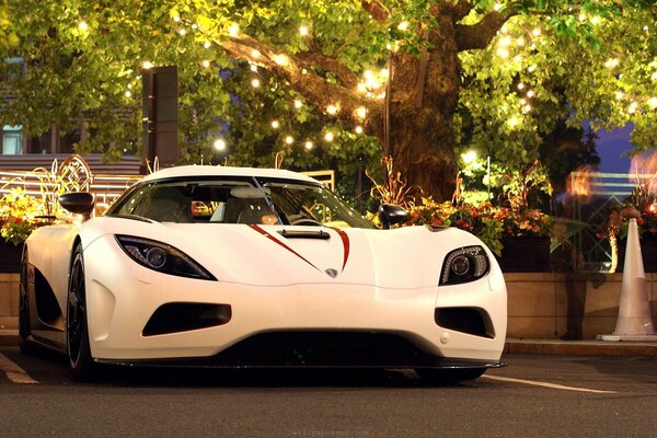 Белый гиперкар Koenigsegg Agera под деревом с гирляндой