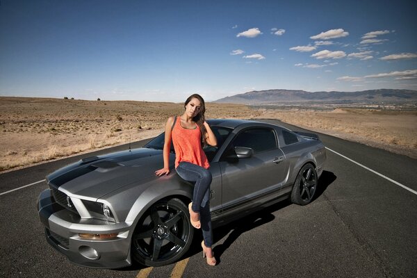 A beautiful girl in an orange top next to a mustang GT 500 in the desert