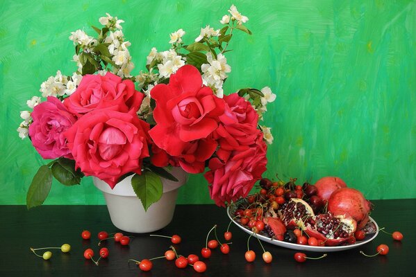 Sur la table se trouve un pot de roses et une assiette de grenades et de cerises
