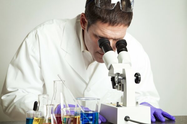 A scientist in a white coat and gloves looks into a microscope