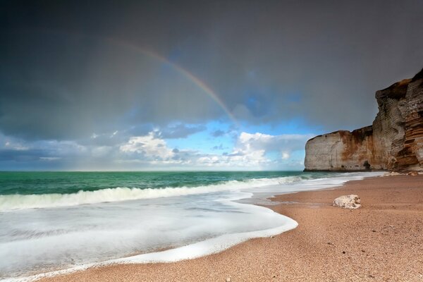 Mar verde, oleaje espumoso y costa de arena con roca en el fondo de un arco iris brumoso